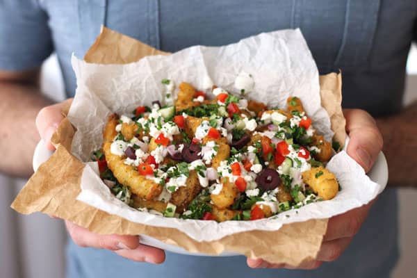 Hands holding a bowl of toaster oven baked fries covered with a tangy yogurt sauce, fresh veggies and feta cheese.