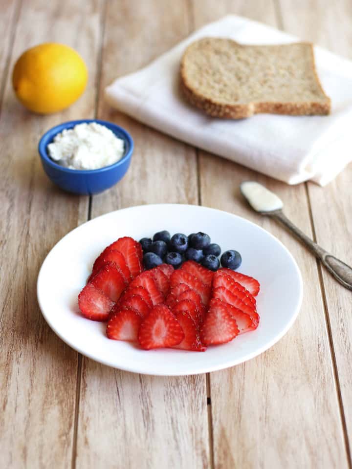 Fresh blueberries and strawberry slices, a small bowl of ricotta, and a piece of bread.