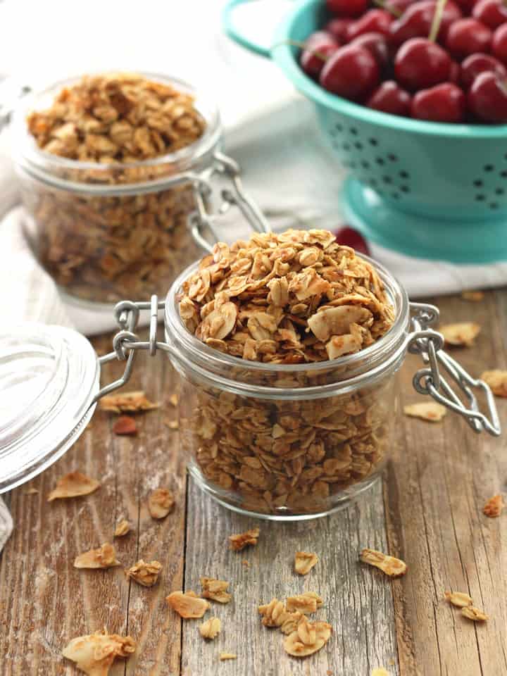 A wooden table with granola in mason jars next to a blue colander full of cherries.