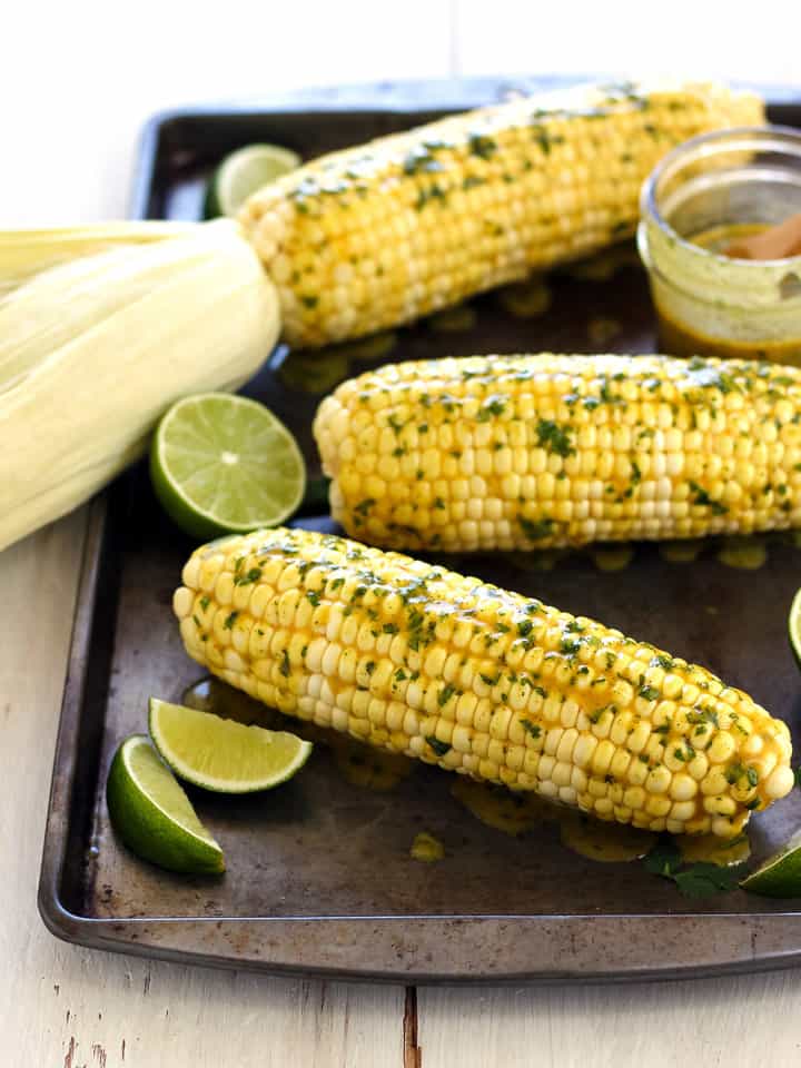 3 baked corn cobs on a sheet pan with lime wedges and seasoned butter.