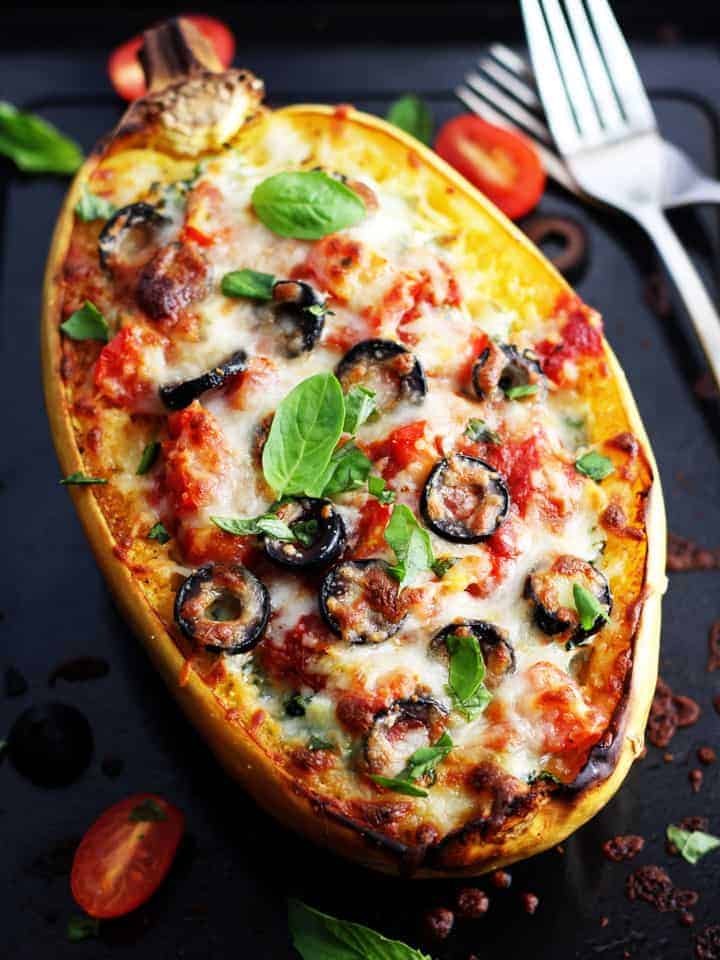 A cheesy spaghetti squash boat topped with olives and tomatoes on a dark roasting pan.