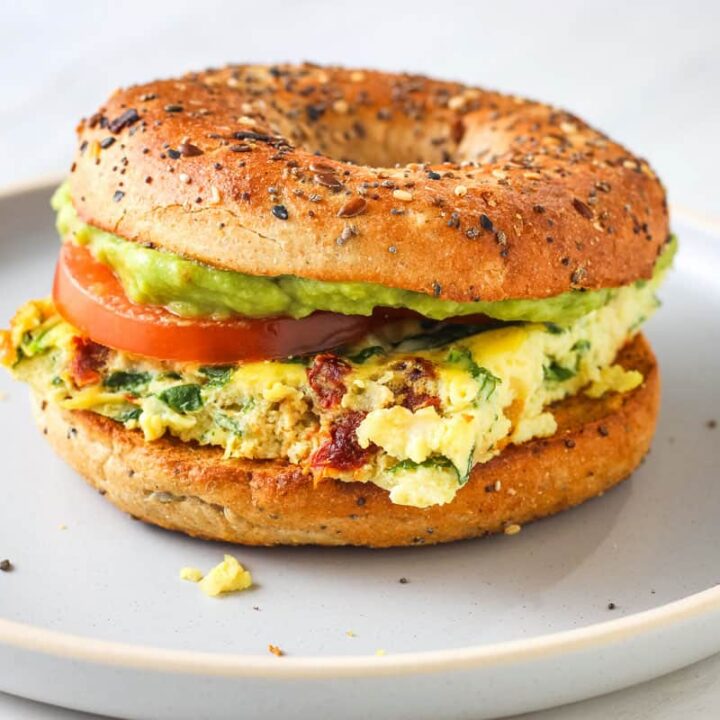 Bagel sandwich layered with eggs, tomato slices, and guacamole on a plate.