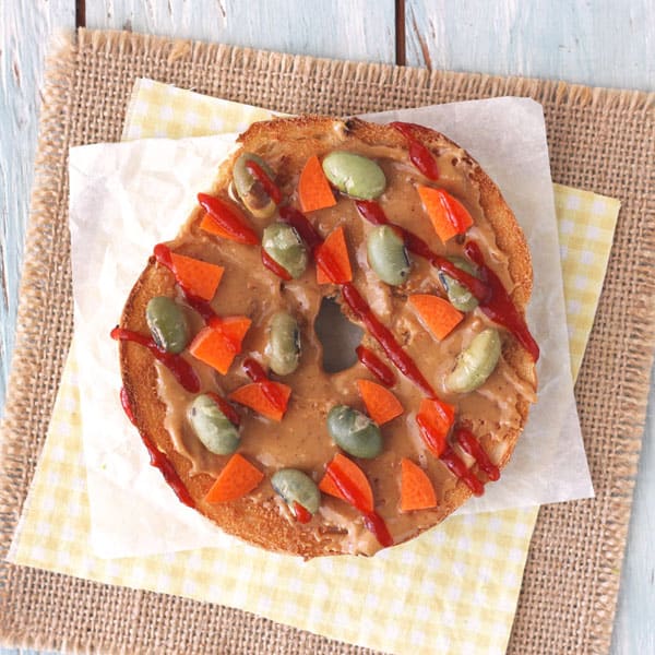 A toasted whole grain bagel topped with peanut butter, carrots, roasted edamame and sriracha sauce.