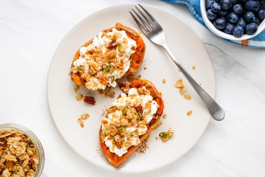 Topped sweet potatoes on a plate next to a jar of granola and bowl of blueberries.