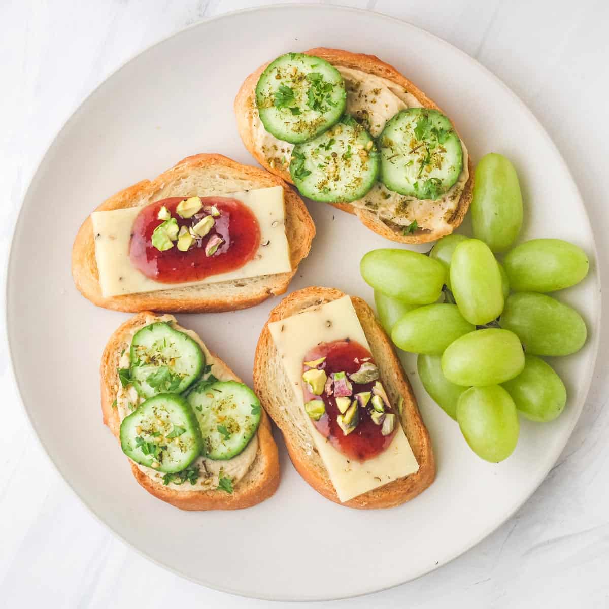 Crostini topped with hummus and cheese on a plate with grapes.