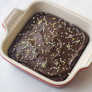 A mini chocolate cake with chocolate frosting and sprinkles in a 5x5 stoneware baking dish.