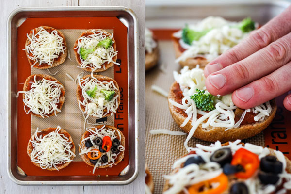 Uncooked pizza bagels on a sheet pan with a hand pressing down onto the toppings.