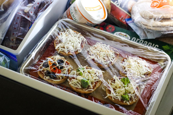Plastic wrap covered pan of pizza bagels inside a freezer.