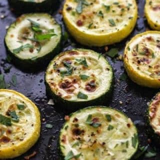 Broiled zucchini and summer squash on a black pan with fresh herbs.