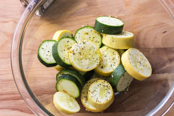 Sliced zucchini in a glass bowl tossed with oil and seasonings.