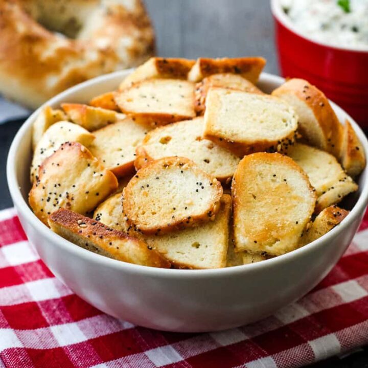 Bowl of bagel crisps on a table with whole bagels and a bowl of dip.