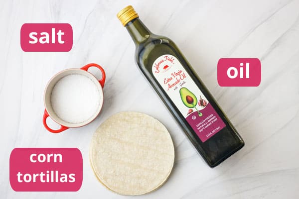 Green bottle of avocado oil, white corn tortillas, and a red ramekin of salt on a table.