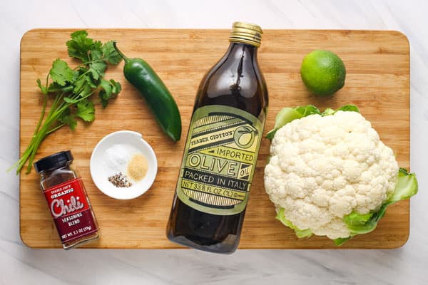 Head of cauliflower, olive oil bottle, cilantro, jalapeno, lime, and seasonings on a cutting board.
