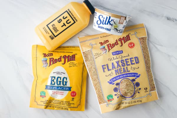 Bags of flax seed and egg replacer, bottle of just egg, and container of yogurt on a table.