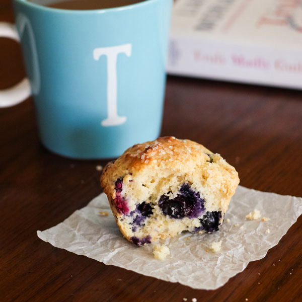 A blueberry muffin, light blue coffee mug and book on a table.