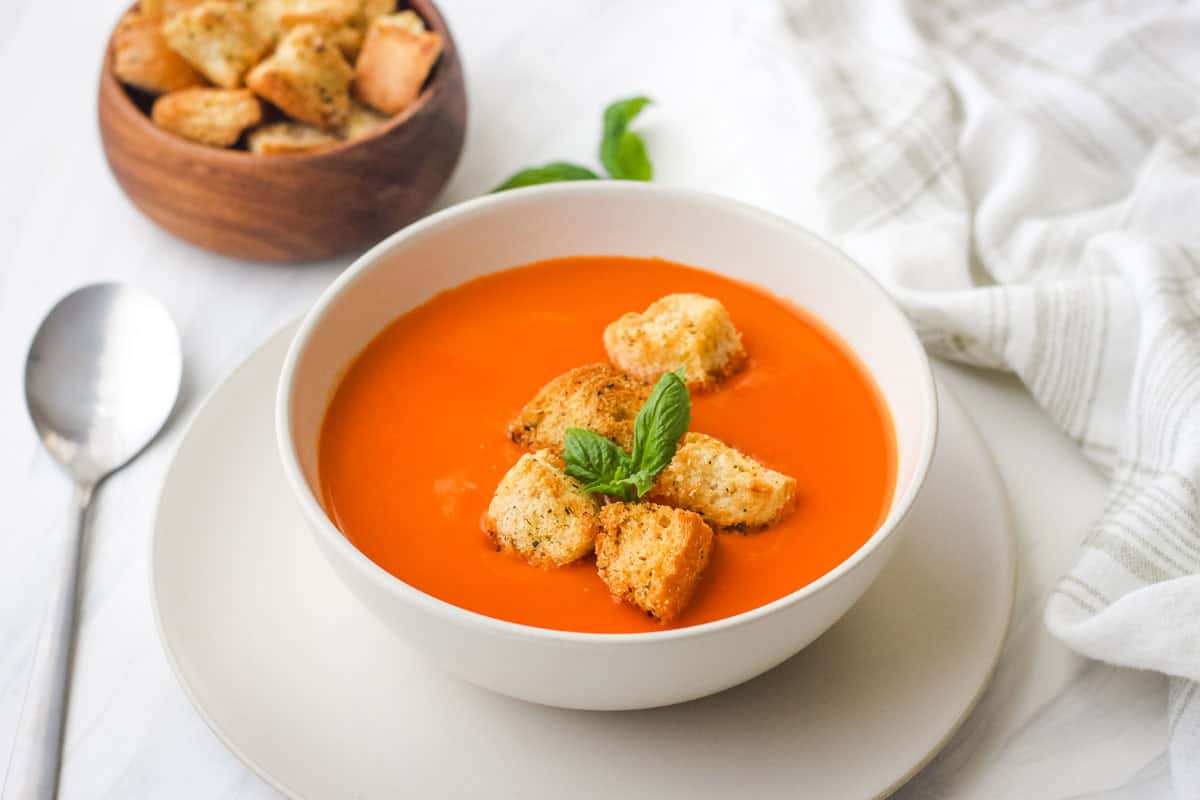 Bowl of red pepper soup topped with croutons and basil leaves.