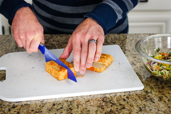 Hands slicing sriracha baked tofu on a cutting board.