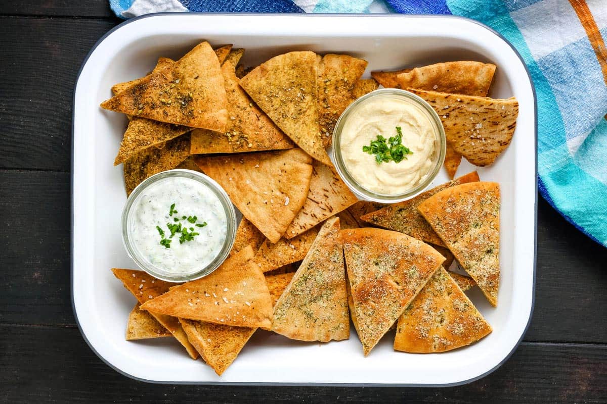 White baking dish with a variety of seasoned pita chips and dips in small mason jars.