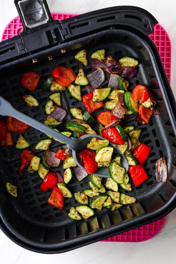 Spoon scooping cooked vegetables in an air fryer basket.