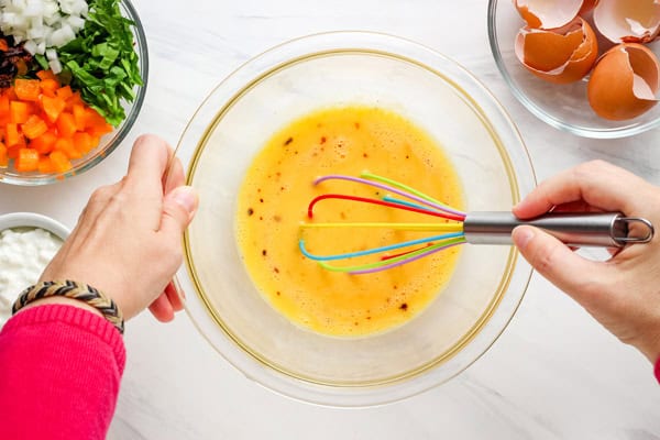 Hands beating eggs in a glass bowl with a whisk.