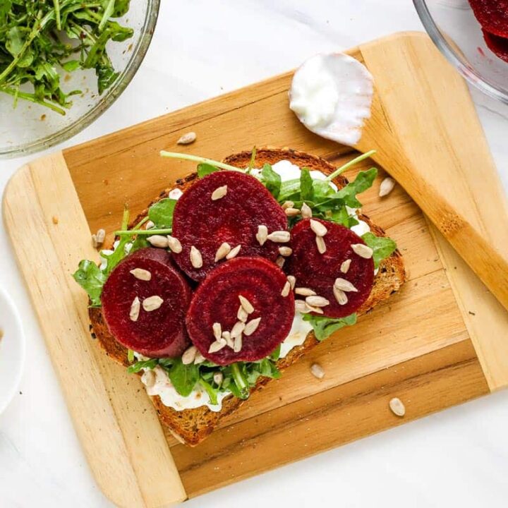 Beet toast on a cutting board with bowls of cottage cheese, arugula salad, and sunflower seeds.