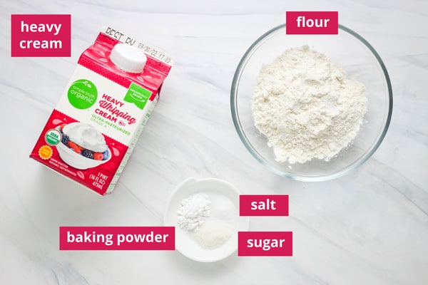 Carton of cream, bowl of flour, and ramekin with salt, sugar, and baking powder arranged on a counter.