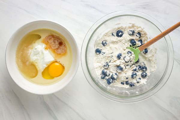 White bowl of wet ingredients next to glass bowl of flour and blueberries.