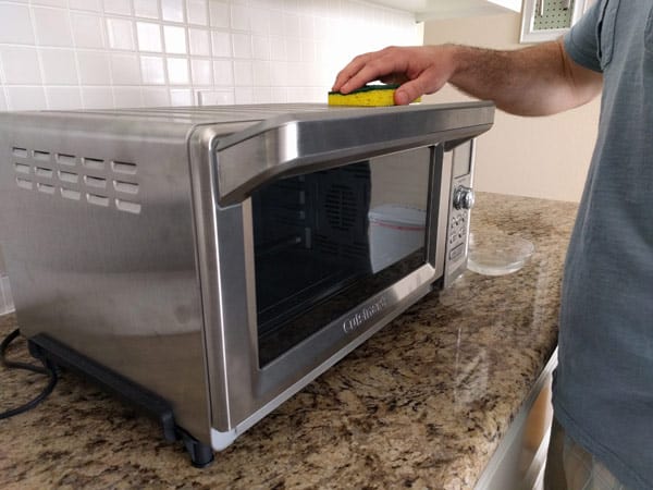 Man wiping down the outside of a toaster oven with a yellow sponge.