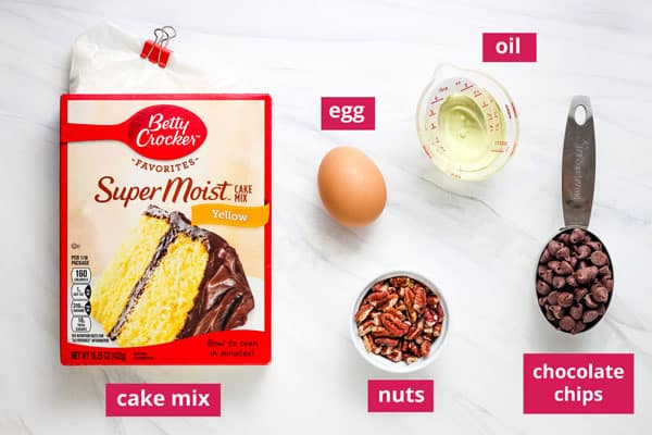 Boxed cake mix, egg, oil. nuts, and measuring cup of chocolate chips arranged on a table and labeled.