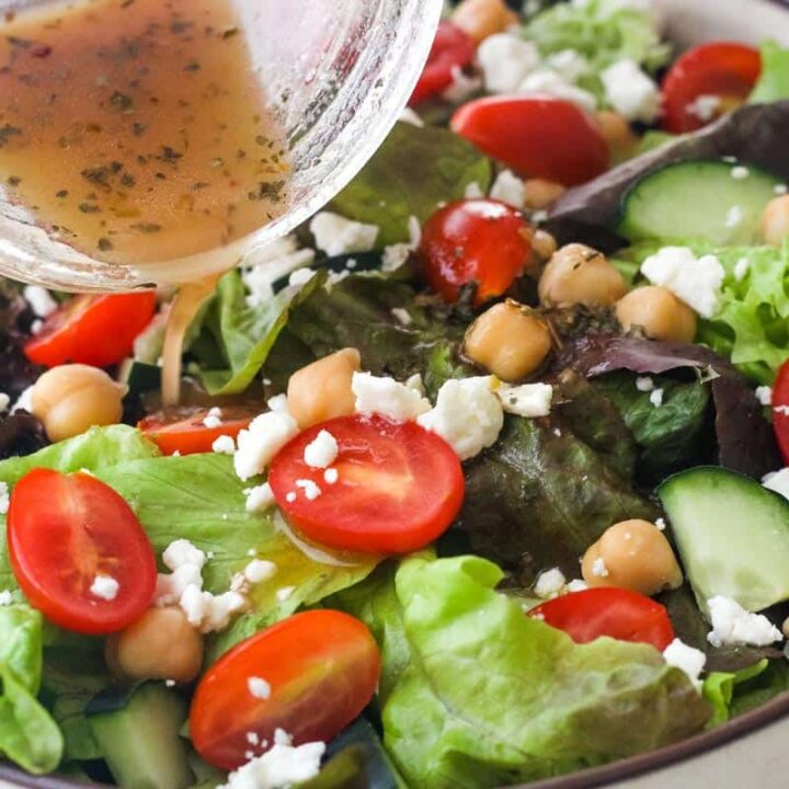 Hand pouring jar of salad dressing over a large green salad.