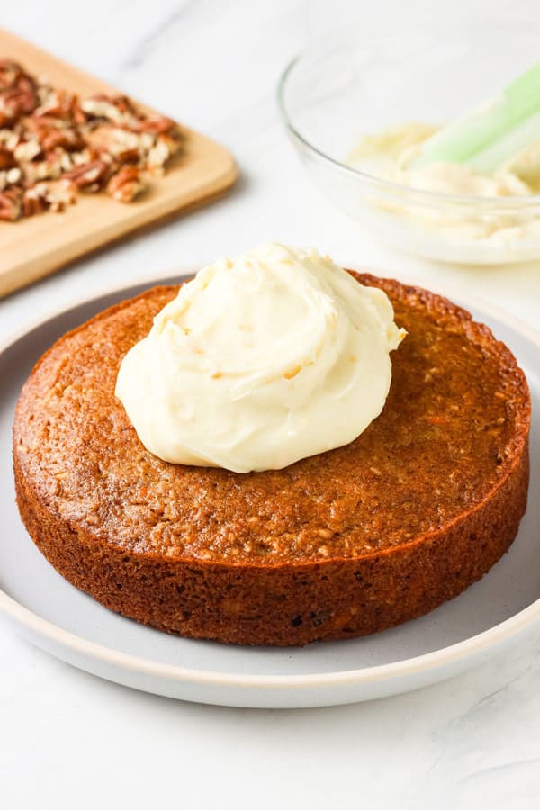 Carrot cake on a plate with a mound of frosting on top next to a cutting board of chopped pecans.