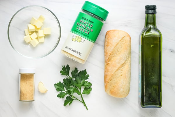 Cubed butter, jar of cheese, fresh parsley, sub roll, bottle of olive oil, jar of garlic powder and garlic clove arranged on a table.