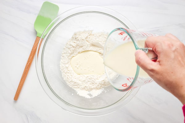 Hand pouring measuring cup of cream over flour in a mixing bowl.