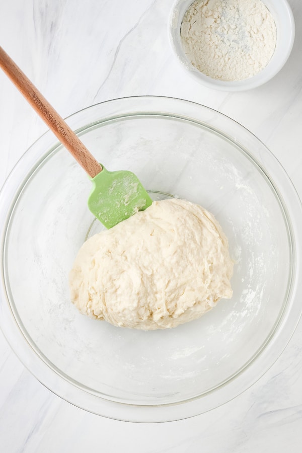 Shaggy sticky dough in a mixing bowl with a rubber spatula and small ramekin of flour.