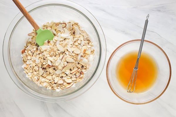 Glass bowl with oats, nuts, and coconut next to a small bowl of liquid with a mini whisk.
