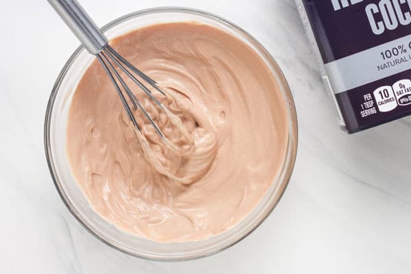 Metal whisk mixing a creamy bowl of mocha yogurt next to a container of cocoa powder.