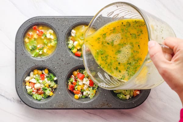 Hand pouring egg mixture from measuring cup over veggies in a muffin tin.