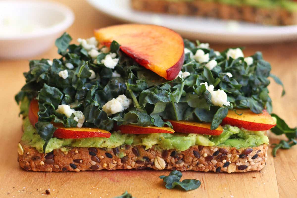 Closeup of toasted bread topped with peach slices, avocado, greens, and crumbled gorgonzola cheese.
