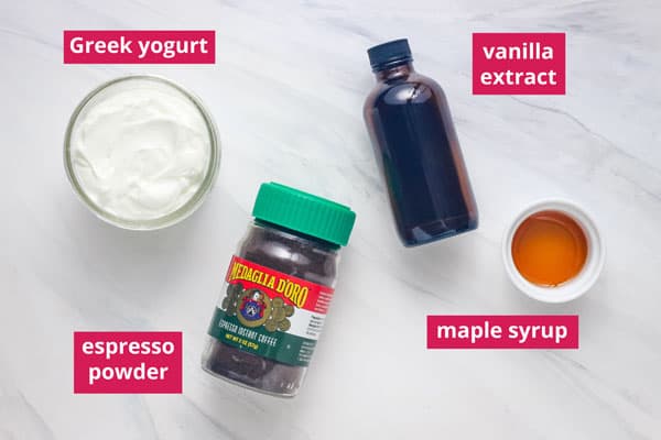 Jar of yogurt and ramekin of maple syrup next to bottles of vanilla extract and espresso powder labeled on a table.