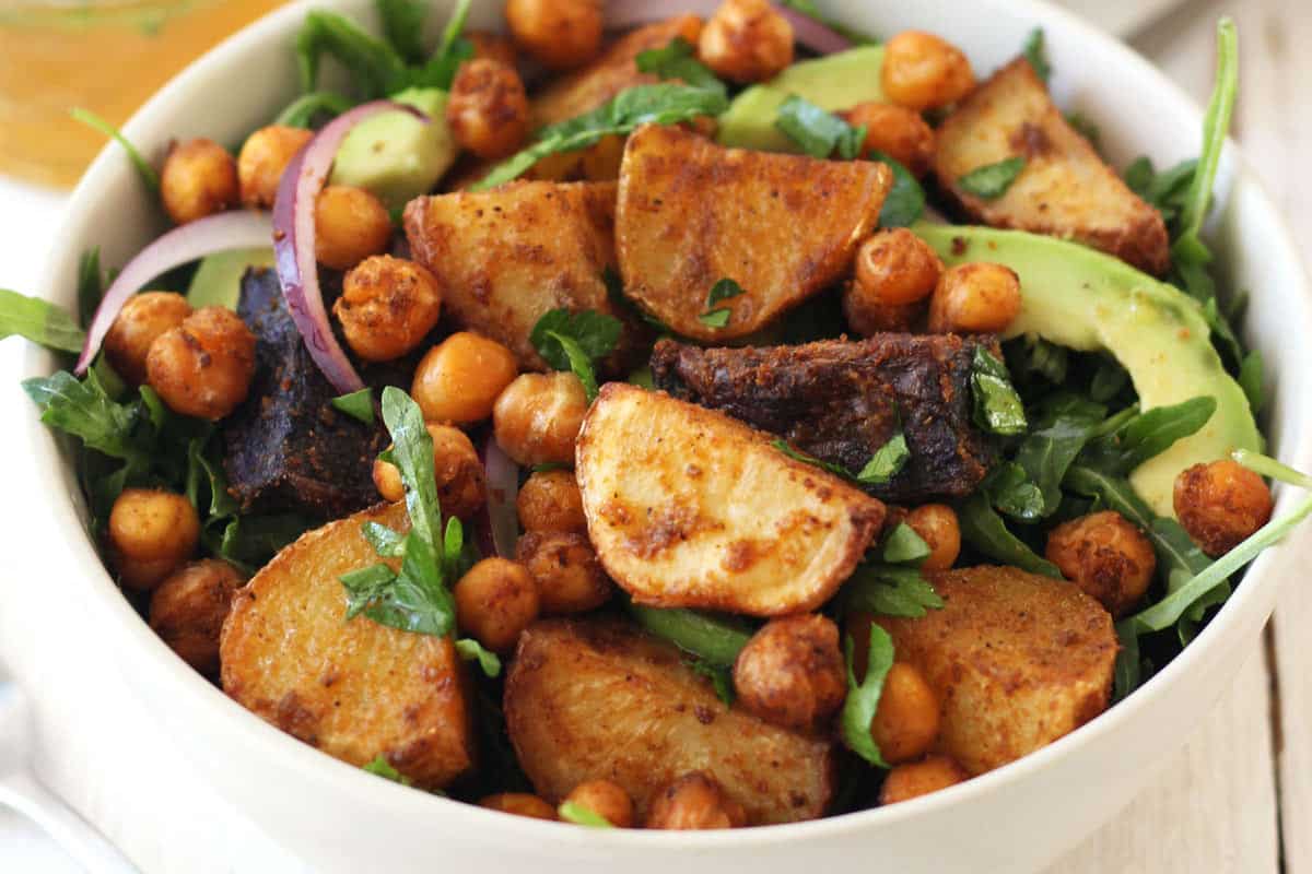 Closeup of roasted chickpeas, potato chunks, and arugula in a white bowl.