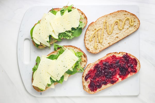 Bread slices on a cutting board topped with cheese, mustard, and cranberry sauce.