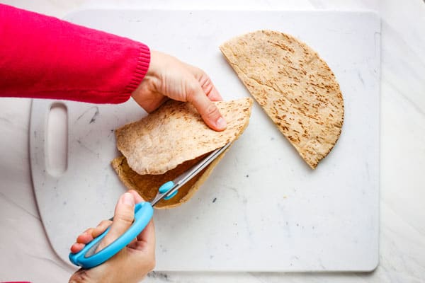 Hands using kitchen scissors to cut pita bread into two layers.
