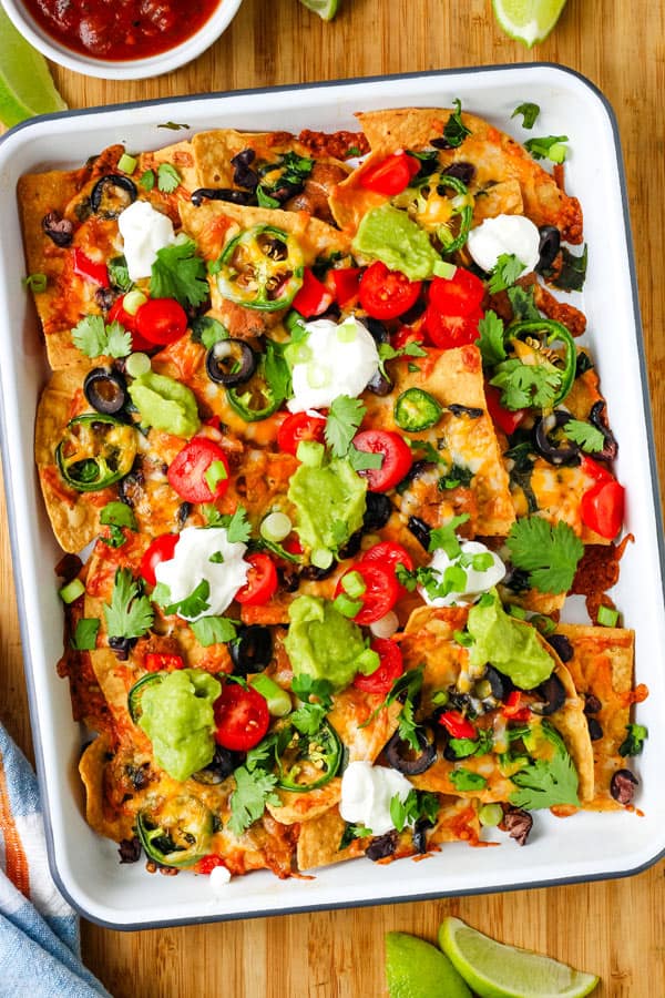 Baked sheet pan nachos on a cutting board with lime wedges and a bowl of salsa.