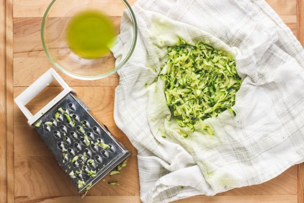 Cutting board with box grater and shredded zucchini in a towel next to a bowl of green liquid.