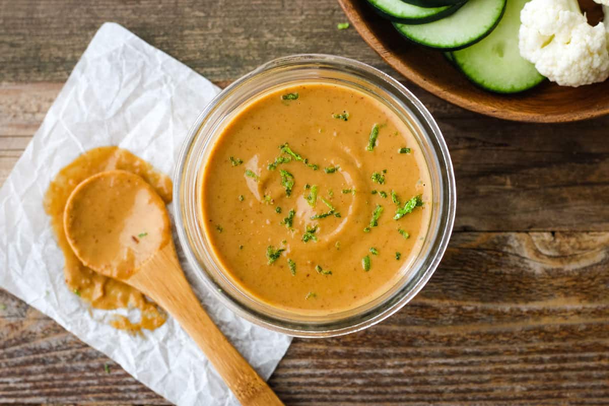 Small jar of peanut sauce with lime zest next to wooden bowl of raw veggies.