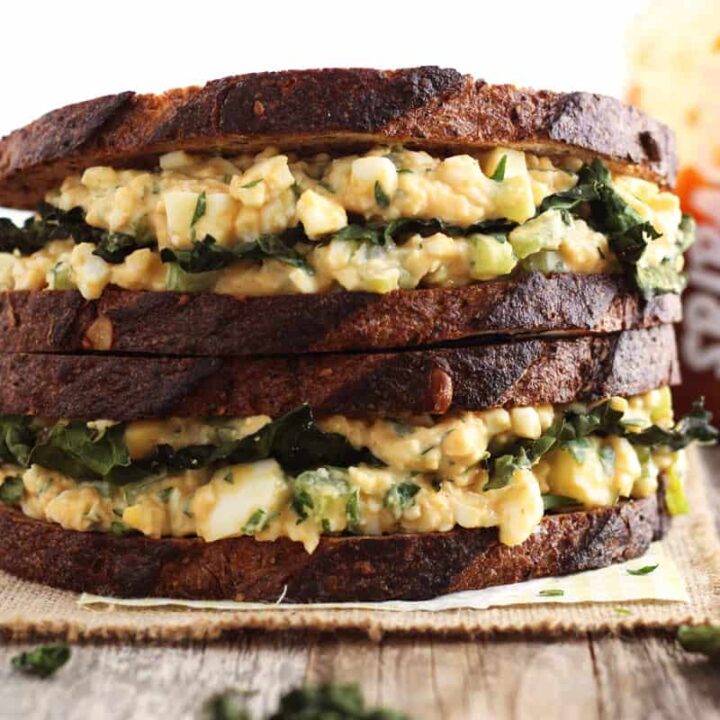 Stacked egg salad sandwiches on whole grain bread, sriracha sauce bottle, and pan of pickle wedges on a table.
