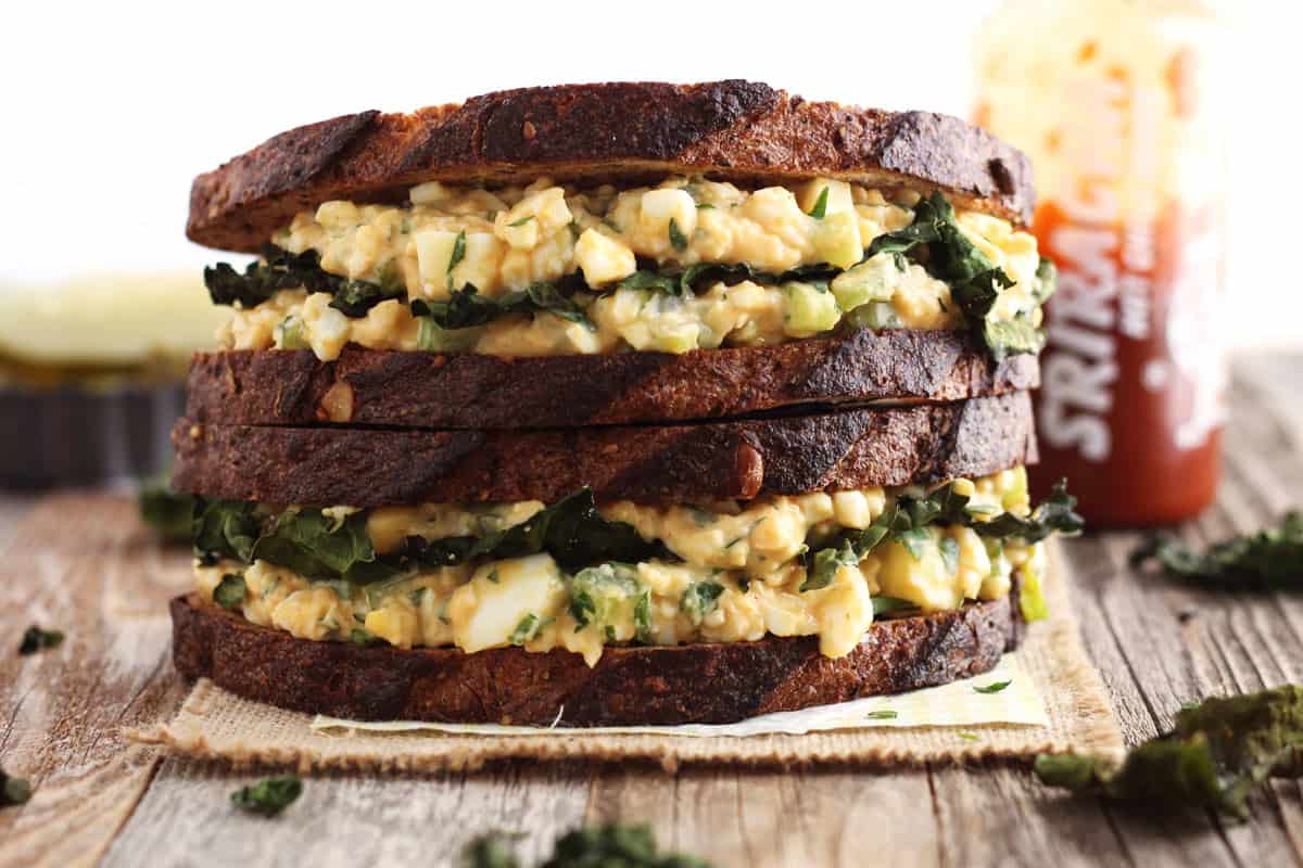 Stacked egg salad sandwiches on whole grain bread, sriracha sauce bottle, and pan of pickle wedges on a table.