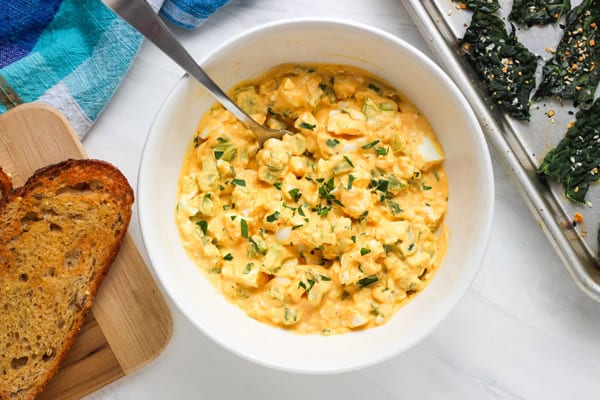 Prepared egg salad in bowl next to toasted bread and a pan of kale chips.