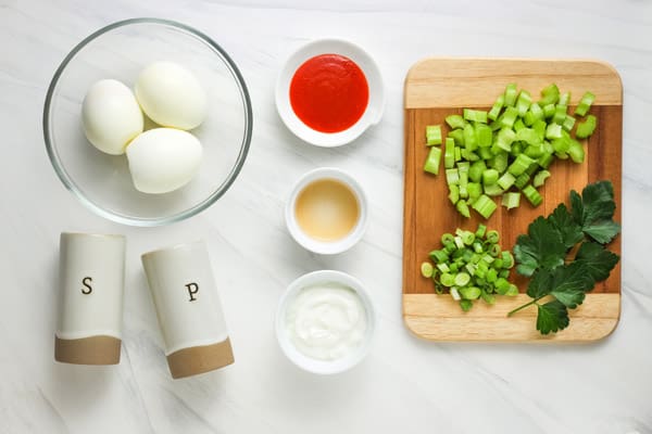 Cutting board with chopped celery, green onion, and parsley. Peeled hard cooked eggs, vinegar, sriracha, and Greek yogurt in bowls arranged on a table.