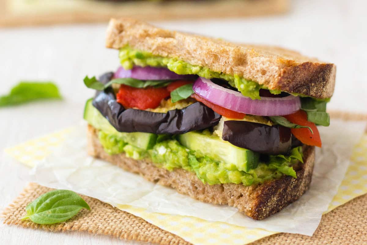 Closeup up of sandwich with mashed avocado and layers of cucumber, eggplant slices, red pepper, basil, and red onions.